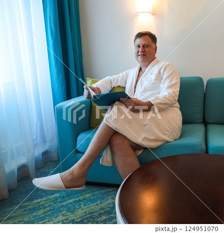 A Man in a Robe Relaxes in His Hotel Room, Engrossed in Reading a Brochure, Showcasing the Comfort and Leisure of Travel, as Well as the Intrigue of Exploring New Destinations 124951070