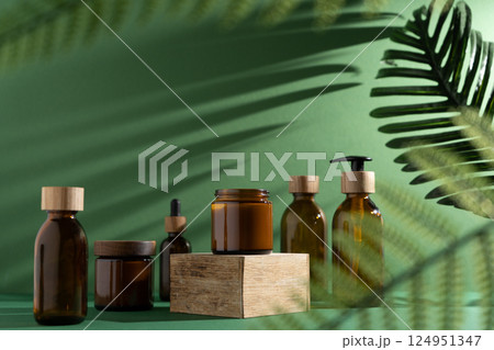 Bottles for cosmetics and cream on a pedestal, green background with leaves and shadow. Brown glass jars with wooden lids. Eco concept. Bottles for cosmetics and cream on a pedestal, green background with leaves and shadow. Brown glass jars with wooden lids. Eco concept. 124951347