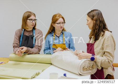 Three female seamstresses fashion designers stand at a table with roll of fabric and negotiate. Three female seamstresses fashion designers stand at a table with roll of fabric and negotiate. 124956517