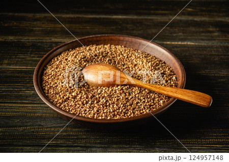 A close-up of a wooden bowl filled with brown buckwheat seeds. A wooden spoon rests on top of the buckwheat grains, creating a rustic and natural atmosphere, perfect for culinary use A close-up of a wooden bowl filled with brown buckwheat seeds. A wooden spoon rests on top of the buckwheat grains, creating a rustic and natural atmosphere, perfect for culinary use 124957148