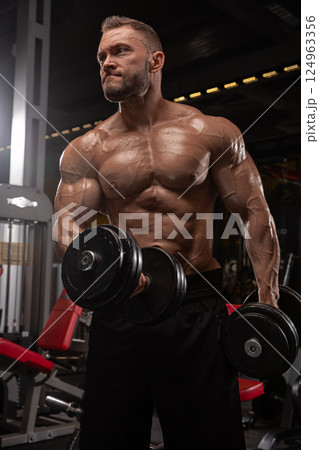 muscular man lifts dumbbells in well equipped gym during evening. He exhibits strong determination as he engages in his workout routine focused on building strength and fitness. 124963356