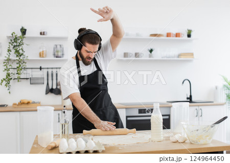 Bearded young adult male wearing black apron and headphones baking in bright kitchen, rolling dough enthusiastically while listening to music. 124964450