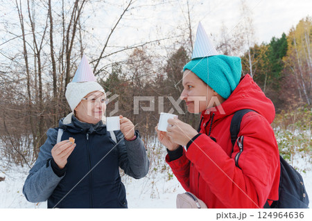 Friends enjoy a cheerful winter day surrounded by snow-covered trees, wearing colorful party hats while sipping warm beverages Friends enjoy a cheerful winter day surrounded by snow-covered trees, wearing colorful party hats while sipping warm beverages 124964636