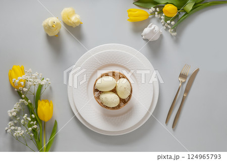 Easter table setting with spring yellow tulips, white plates, bunny, eggs on gray background. Happy Easter celebration. Copy space. View from above. Easter table setting with spring yellow tulips, white plates, bunny, eggs on gray background. Happy Easter celebration. Copy space. View from above. 124965793