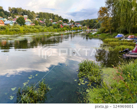 Village lake with houses and boats on the shore, banks overgrown with reeds, summer rural calm landscape Village lake with houses and boats on the shore, banks overgrown with reeds, summer rural calm landscape 124969801