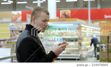 Man carefully reads labels in crowded supermarket, selecting nutritious items. Daily ritual of shopper navigating diverse choices. A man studies the label on a bottle of sauce Man carefully reads labels in crowded supermarket, selecting nutritious items. Daily ritual of shopper navigating diverse choices. A man studies the label on a bottle of sauce 124979005