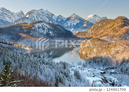 Snowy Mountain Landscape with Iconic Castle Overlooking Serene Lake and Alpine Peaks in Ethereal Winter Wonderland Bathed in Soft Dawn Light 124981709