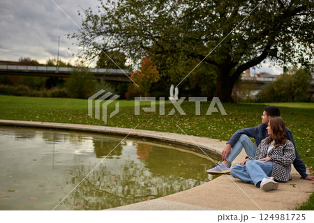Romantic young couple relaxing by the pond in autumn park, wearing cozy casual outfits, enjoying a peaceful moment together. 124981725