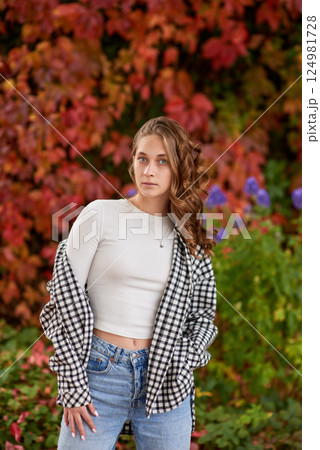 Autumn Fashion Portrait: Young Woman in Casual Outfit Posing Against Vibrant Red Ivy Wall in Urban Outdoor Setting. Stylish Seasonal Style Showcases Warm Earthy Tones and Cozy Fall Atmosphere. 124981728