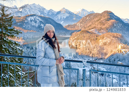 Winter Wonderland Serenity: Woman in Grey Puffer Jacket Admiring Snowy Mountain Landscape with Golden Sunlight and Picturesque Village from Balcony 124981833