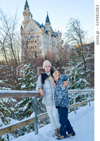 Mother and Child Exploring Snowy Winter Castle Wonderland on Wooden Bridge Against Blue Sky with Historic Architecture and Snow Covered Landscape 124981863