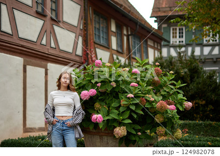 Young woman posing with hydrangeas in a European garden, surrounded by half-timbered houses and autumn foliage, expressing joy and elegance. 124982078