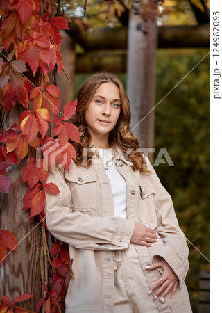 Smiling young woman in beige casual outfit leaning on wooden pillar with red autumn leaves in scenic garden pergola – warm and cozy fall portrait with natural background 124982093