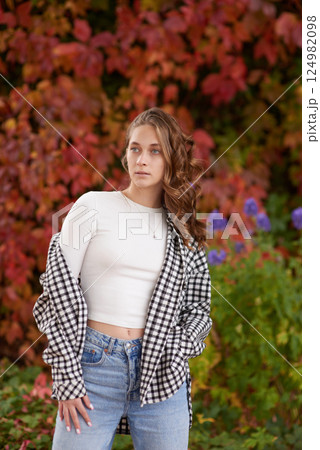 Young woman in casual autumn outfit posing against vibrant red ivy wall, stylish fashion portrait in warm seasonal tones. 124982098