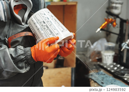 Worker in protective gear holds a paint container, preparing for spray painting in a workshop during the day Worker in protective gear holds a paint container, preparing for spray painting in a workshop during the day 124983592