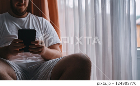 Casual man at home using smartphone while sitting by window with light curtains in cozy indoor 124985770