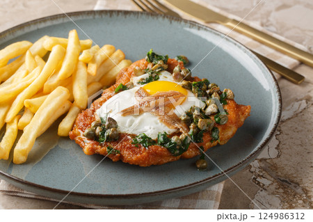 Delicious breaded schnitzel with anchovies, fried egg and capers parsley sauce on top served with french fries close-up on a plate. Horizontal 124986312