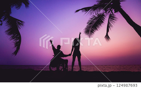 Disabled man in a wheelchair with his wife on the beach. Silhouettes at sunset Disabled man in a wheelchair with his wife on the beach. Silhouettes at sunset 124987693