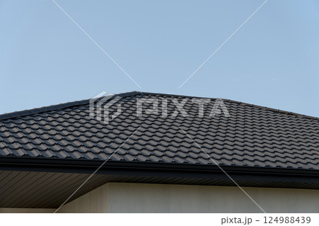 Dark tiled roof covers a residential home, highlighting the craftsmanship in roofing construction against a cloudless background Dark tiled roof covers a residential home, highlighting the craftsmanship in roofing construction against a cloudless background 124988439