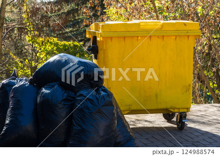 Bright yellow garbage bin stands next to piled black trash bags under clear blue skies in a lush green park setting Bright yellow garbage bin stands next to piled black trash bags under clear blue skies in a lush green park setting 124988574