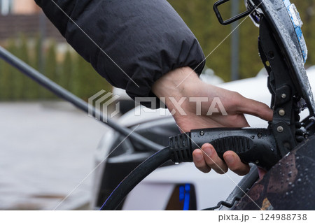 A person connects a charging cable to an electric vehicle at a charging station while it drizzles and overcast skies loom overhead 124988738