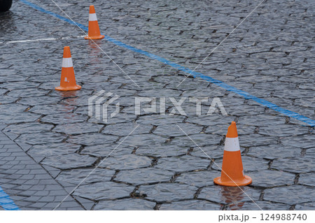 Orange traffic cones sit on a wet cobblestone surface, marking no parking zones and creating safety measures in rainy conditions Orange traffic cones sit on a wet cobblestone surface, marking no parking zones and creating safety measures in rainy conditions 124988740
