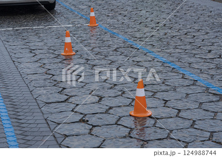 Wet pavement in an urban parking lot features orange cones spaced evenly, indicating reserved spaces during a rainy day Wet pavement in an urban parking lot features orange cones spaced evenly, indicating reserved spaces during a rainy day 124988744