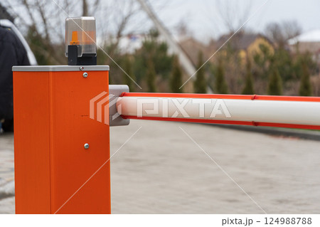A bright orange barrier gate stands at the entrance of a parking lot, with a flashing light indicating restricted access to vehicles 124988788