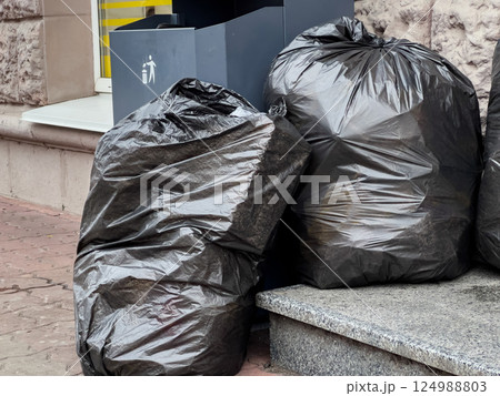 Several black trash bags sit next to a waste bin on a busy urban sidewalk, showing signs of recent litter collection efforts 124988803