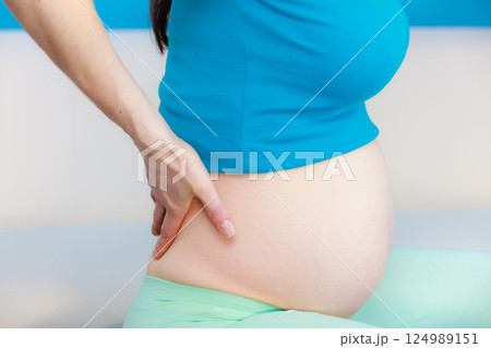 Pregnant woman sitting on couch holds hand on her back Pregnant woman sitting on couch holds hand on her back 124989151