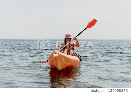 Kayaking Woman Paddle Ocean - A woman wearing a pink hat kayaks on the ocean, paddling with an orange paddle. Kayaking Woman Paddle Ocean - A woman wearing a pink hat kayaks on the ocean, paddling with an orange paddle. 124990376