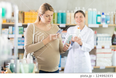 Pregnant woman in pharmacy scans QR code on antifungal cream, pharmacist offer similar product in background. 124992835