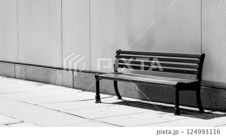 Minimalist image of a black metal bench with a wooden seat against a textured white wall, creating striking contrast. Long shadow adds depth and drama to the serene composition 124993116