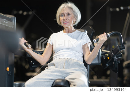 Elderly woman performing biceps curls on exercise machine in gym 124993164