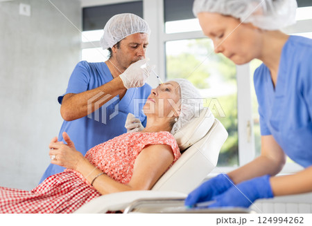 Male doctor with help of nurse performs rejuvenation procedure for elderly female in forehead area 124994262