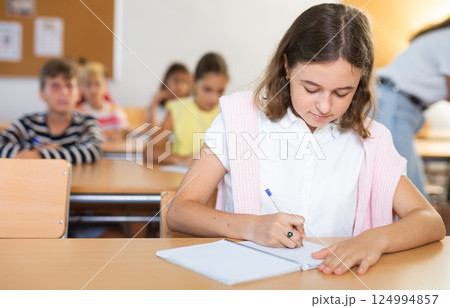 Assiduous focused tween girl sitting with pen and notebook at lesson Assiduous focused tween girl sitting with pen and notebook at lesson 124994857