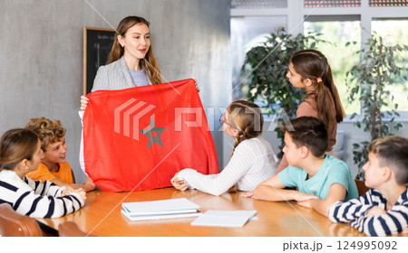 Group of curious preteen learners and young female teacher with flag of Morocco in classroom of academy Group of curious preteen learners and young female teacher with flag of Morocco in classroom of academy 124995092