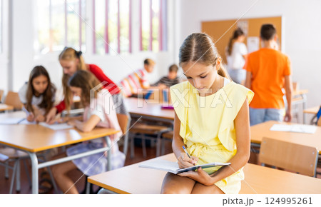 Portrait of schoolgirl with notebook in her hands who sits on desk in school class 124995261