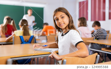 Schoolgirl sits half-turned at desk in classroom at lesson in elementary school Schoolgirl sits half-turned at desk in classroom at lesson in elementary school 124995388