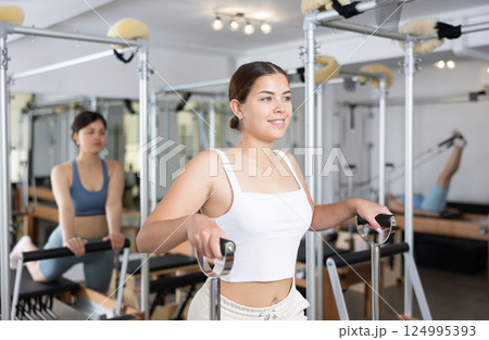 Young positive woman stretches her body muscles while sitting on a special chair in Pilates gym 124995393
