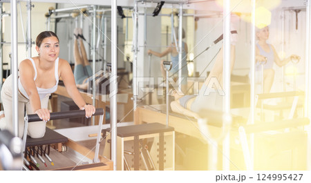 Positive young female in sportswear training twist rotation while using pilates performer bed in gym Positive young female in sportswear training twist rotation while using pilates performer bed in gym 124995427