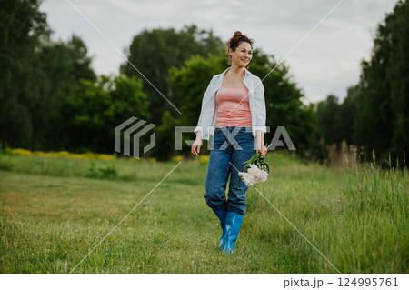 A woman walks with a bouquet of peonies in the backyard of a country house on a summer day. Woman enjoying slow life, nature and harmony in a rustic setting, relaxation 124995761