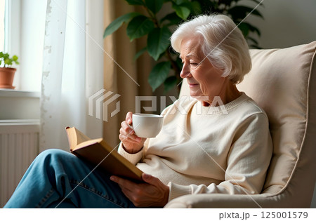 Elderly woman with glasses sipping coffee while reading book in cozy living room 125001579
