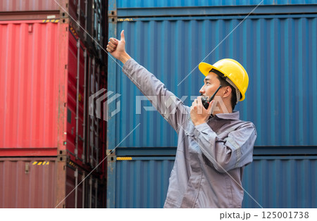 Male dock worker in yellow hard hat and grey uniform gives thumbs up signal while speaking on walkie talkie at shipping container yard Male dock worker in yellow hard hat and grey uniform gives thumbs up signal while speaking on walkie talkie at shipping container yard 125001738