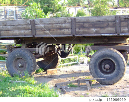 A rustic wooden wagon stands on a gravel surface in a yard, showcasing its robust wheels and structure. Surrounding greenery and buildings create a charming countryside atmosphere. 125006969