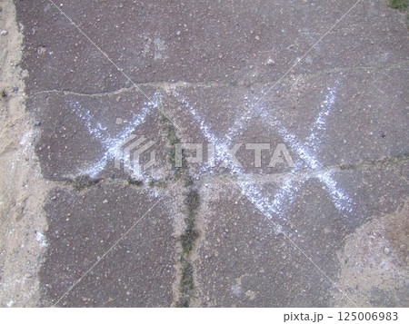 White chalk-like markings appear on a rough asphalt surface. The markings form two crossed 'X' shapes, suggesting a potential point of interest for maintenance or construction work. 125006983