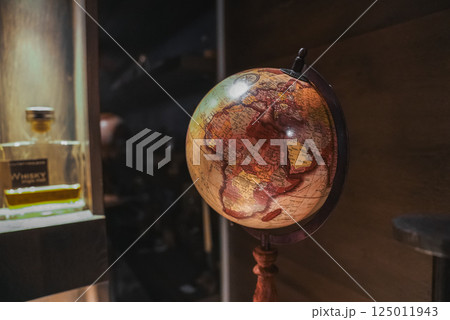 A vintage style globe on a wooden stand displays Europe, Africa, and the Middle East. A whiskey bottle sits on a shelf in a cozy, elegant interior. 125011943