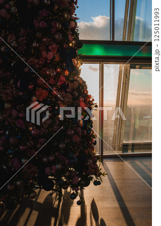 A decorated Christmas tree with ornaments and flowers stands by a window, revealing London's skyline with The Shard at sunset, casting warm shadows. 125011993