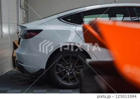A close up view of a white Tesla Model Y's rear side in a garage with a checkered floor. The scene includes a blurred orange car in the foreground. 125011994
