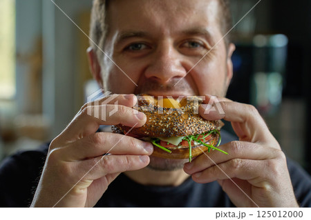 Man takes big bite of seeded bagel filled with egg, cheese, tomato and greens. Hungry man eats sandwich at kitchen, close up 125012000